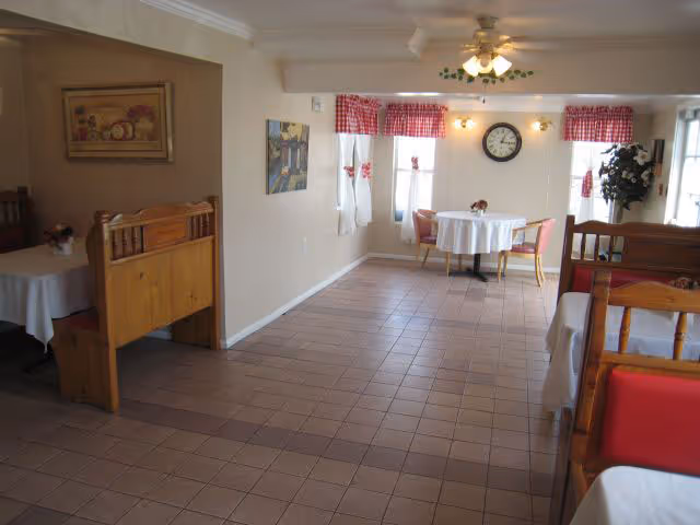 Small dining room with tiled floor, round tables and wooden booths, red-check curtains, a wall clock and ceiling fan.