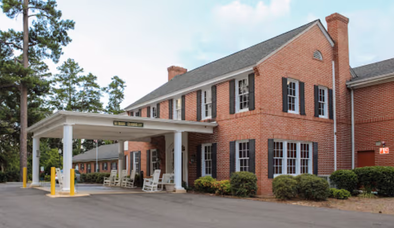 Exterior view of a two-story brick building with black window shutters and a covered entrance with white columns. There are rocking chairs under the covered area and some bushes along the building. Tall trees are visible in the background.