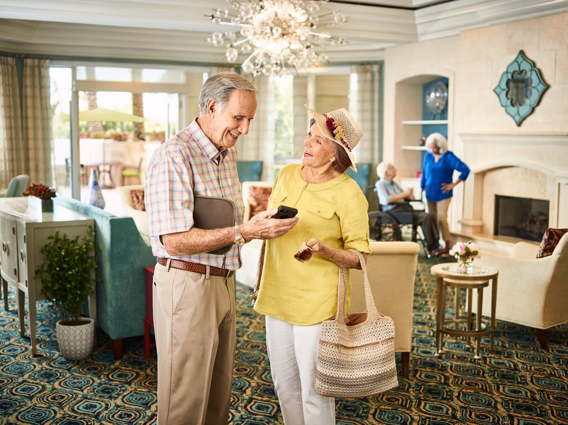 Two elderly people smiling and looking at a smartphone in a well-lit living room with a fireplace, patterned carpet, and comfortable seating. In the background, another elderly person in a wheelchair and a standing woman are conversing near the fireplace.
