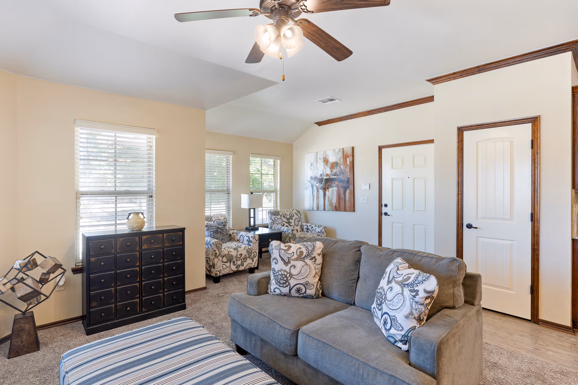 A bright living room with a gray sofa adorned with patterned pillows, a striped ottoman, two floral armchairs near windows with blinds, a black chest of drawers, a modern lamp on a side table, a ceiling fan with lights, and a painting on the wall next to two white doors with wooden trim.