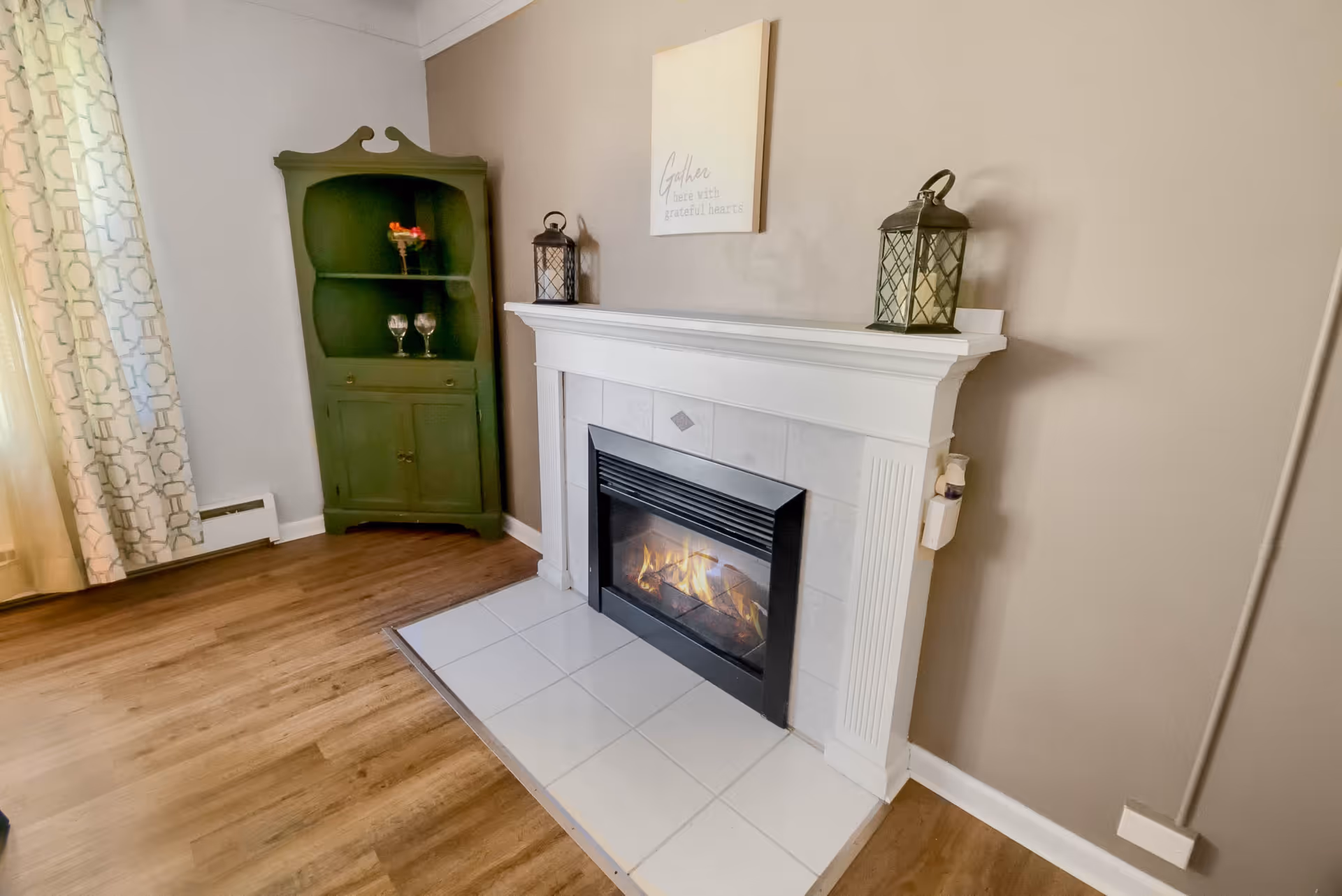 A cozy interior corner featuring a lit fireplace with a white mantel, two decorative lanterns on top, and a small framed sign above it. To the left of the fireplace is a green corner cabinet with glassware and a small flower arrangement. Light-colored curtains with a geometric pattern hang on the window, and the floor is wood with a white tiled hearth in front of the fireplace.