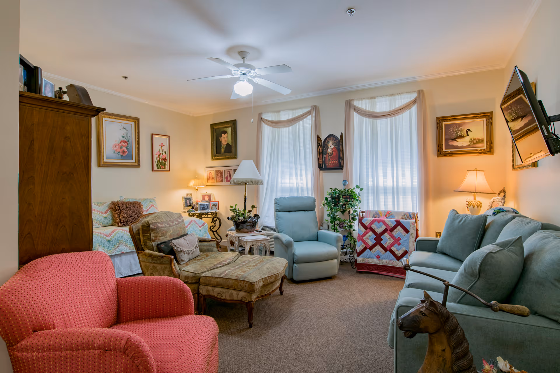 A cozy living room in a senior living facility with a red armchair, a patterned chaise lounge, a light blue recliner, and a blue sofa. The room has beige walls adorned with framed paintings and photographs. Two large windows with sheer white curtains allow natural light to fill the space. There are several lamps providing warm lighting, a ceiling fan with a light, and decorative plants and quilts adding a homey touch.