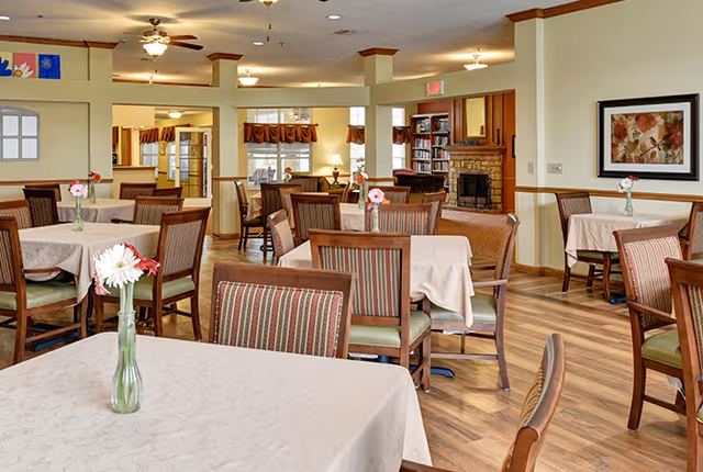 A dining room with multiple tables covered with beige tablecloths, each table having a small vase with flowers. The room has wooden chairs with striped cushions, hardwood flooring, and a cozy fireplace in the background. There are windows with valances and framed artwork on the walls.