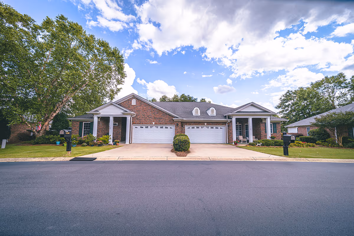 Front exterior view of a single-story brick residential building with two garages, a driveway, green lawns, trees, and a partly cloudy blue sky.