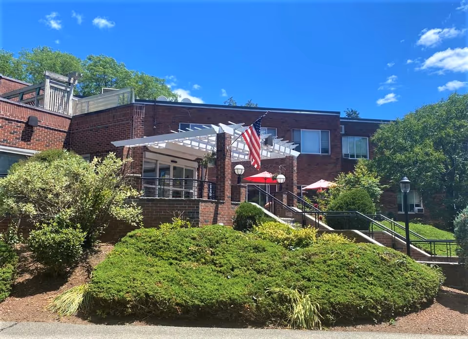 Front exterior of a brick senior living building with a white pergola, an American flag, stairs, patio umbrellas and landscaped greenery under a blue sky.
