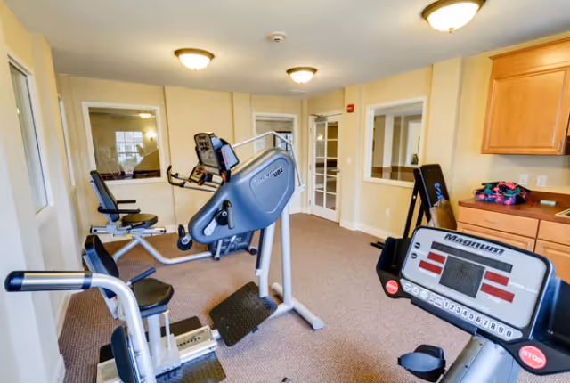 A small fitness room with exercise equipment including a recumbent bike, a seated stepper, and a treadmill. The room has beige walls, carpeted floor, ceiling lights, and windows looking into adjacent rooms. There are wooden cabinets and a countertop on the right side.