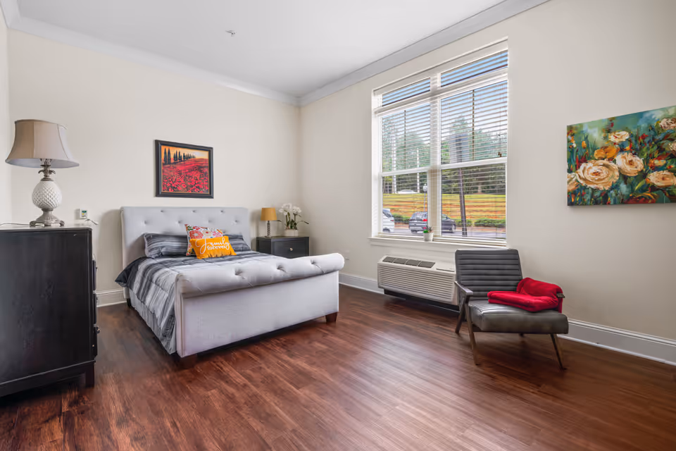 A bright bedroom with a large window letting in natural light. The room features a gray upholstered bed with pillows, a black dresser with a lamp, a black nightstand with a small lamp and flowers, a gray chair with a red blanket, and two colorful paintings on the walls. The floor is wooden, and there is an air conditioning unit below the window.