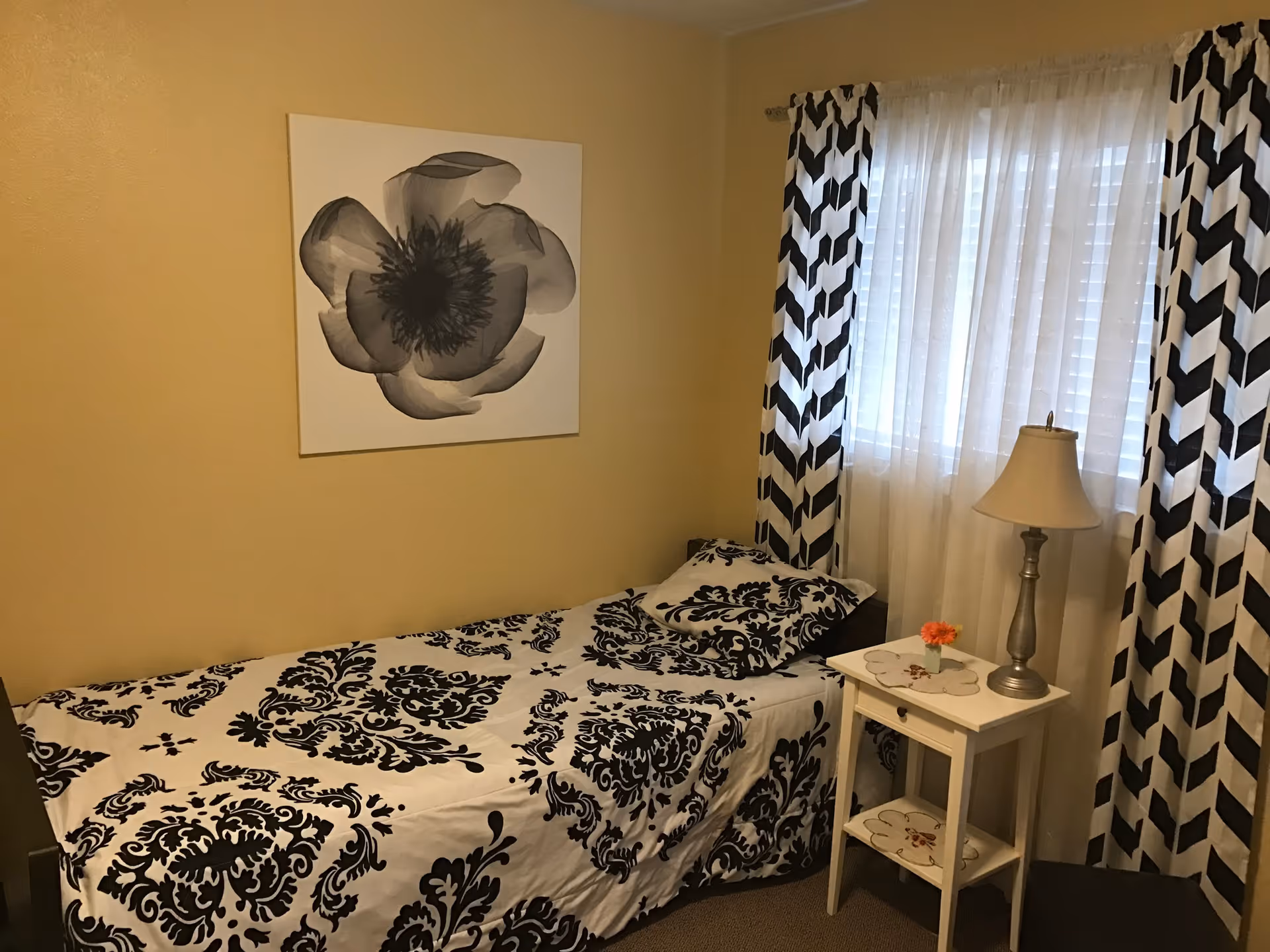 Small single bedroom with black-and-white patterned bedding and curtains, a nightstand with a lamp and a floral wall print.