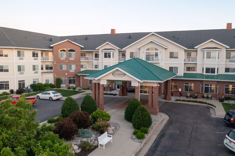 Front exterior of a multi-story senior living building with a green-roofed porte-cochere and landscaped entrance.