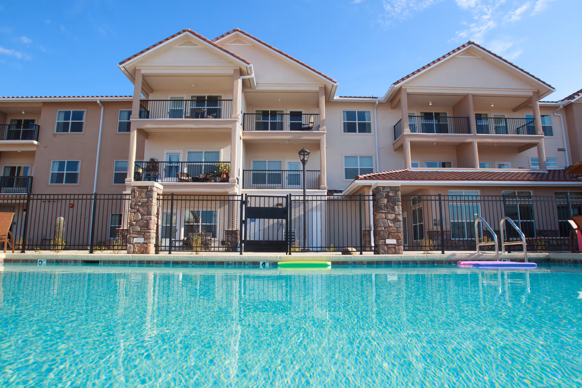 View of a three-story residential building with balconies overlooking a clear blue swimming pool under a bright sky.