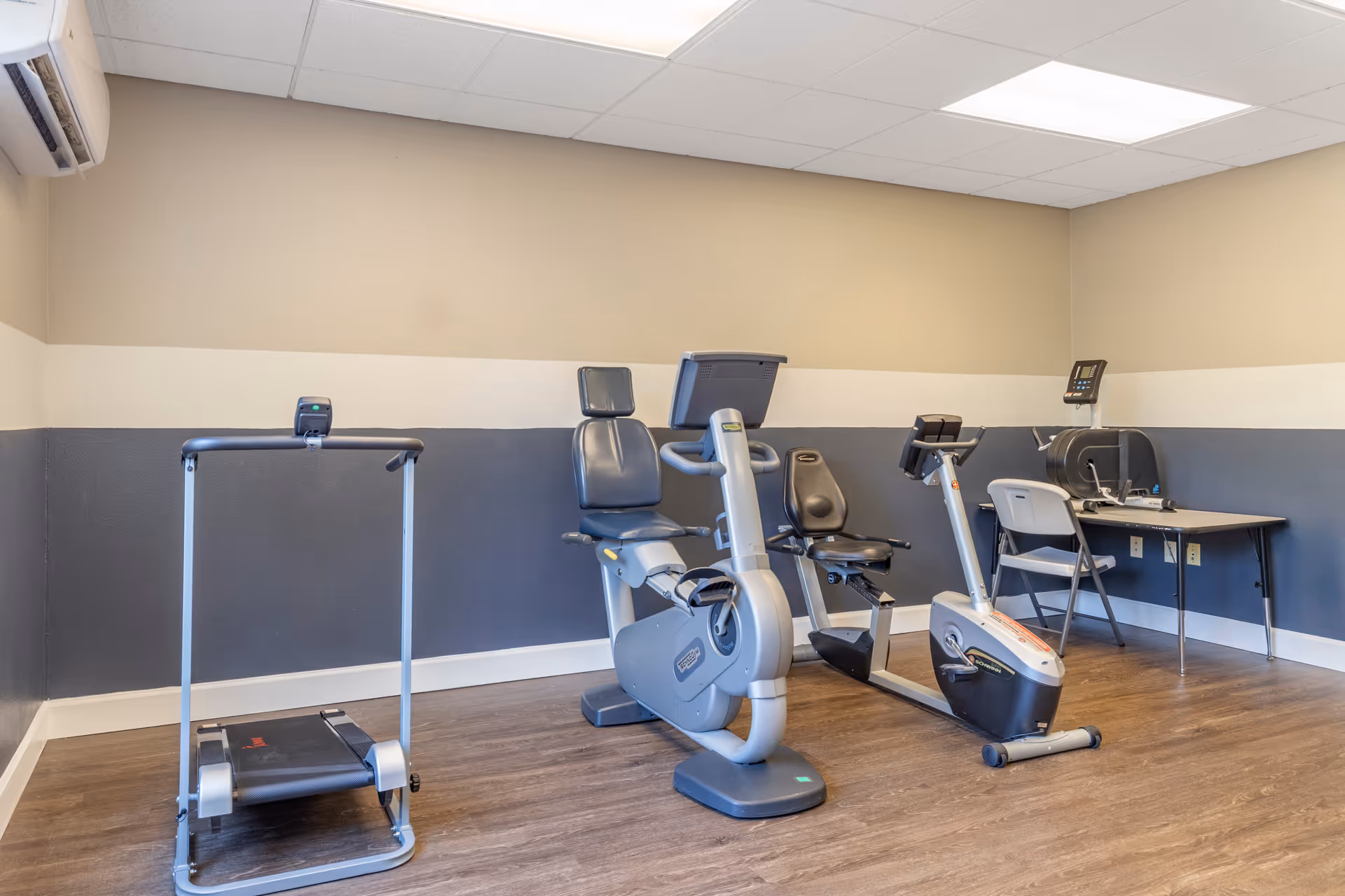 Small exercise room with a treadmill, recumbent and upright stationary bikes, and a table and chair against two-tone walls.