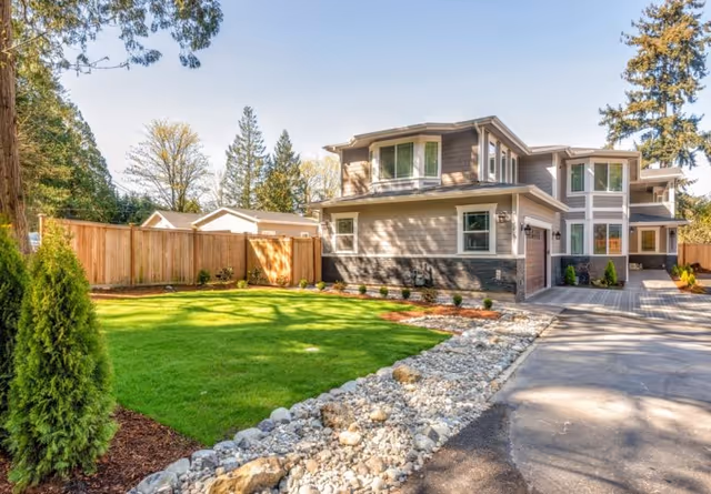 A modern two-story senior care home with gray siding and large windows, surrounded by a well-maintained lawn, small shrubs, and a wooden fence. The driveway is paved and bordered by a rock garden, with tall trees in the background under a clear blue sky.
