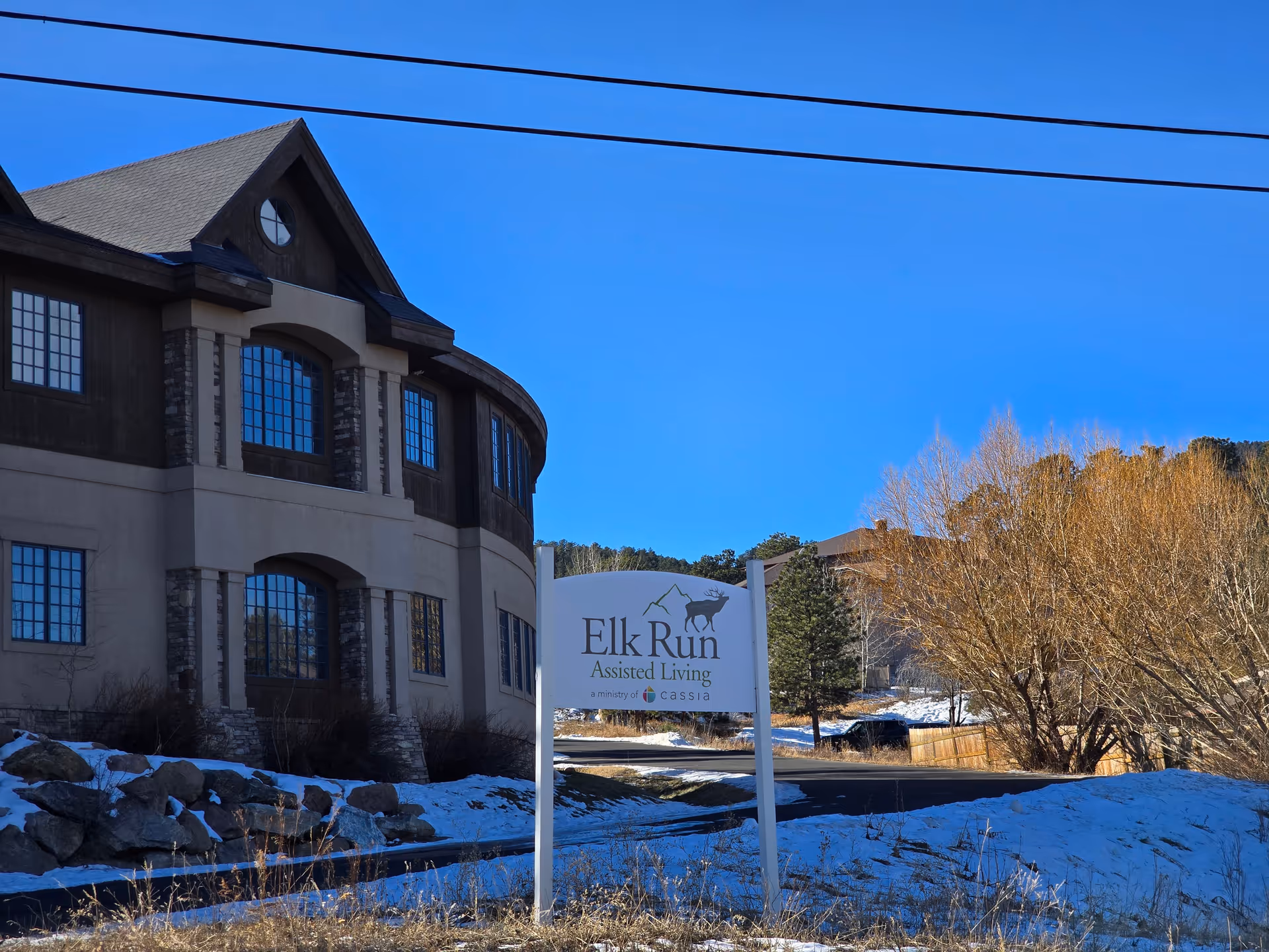 Exterior view of Elk Run Assisted Living facility with a large building on the left and a sign in the foreground displaying the facility's name. Snow covers the ground and trees are visible in the background under a clear blue sky.