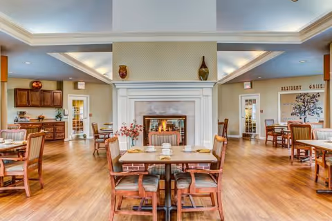 A spacious dining area with wooden floors and multiple tables and chairs arranged neatly. In the center, there is a fireplace with a fire burning, flanked by decorative vases on the mantel. The room has soft lighting with a coffered ceiling and light-colored walls. There are doors and cabinets visible in the background, and a wall with the words 'Resident Gallery' and a decorative tree design.