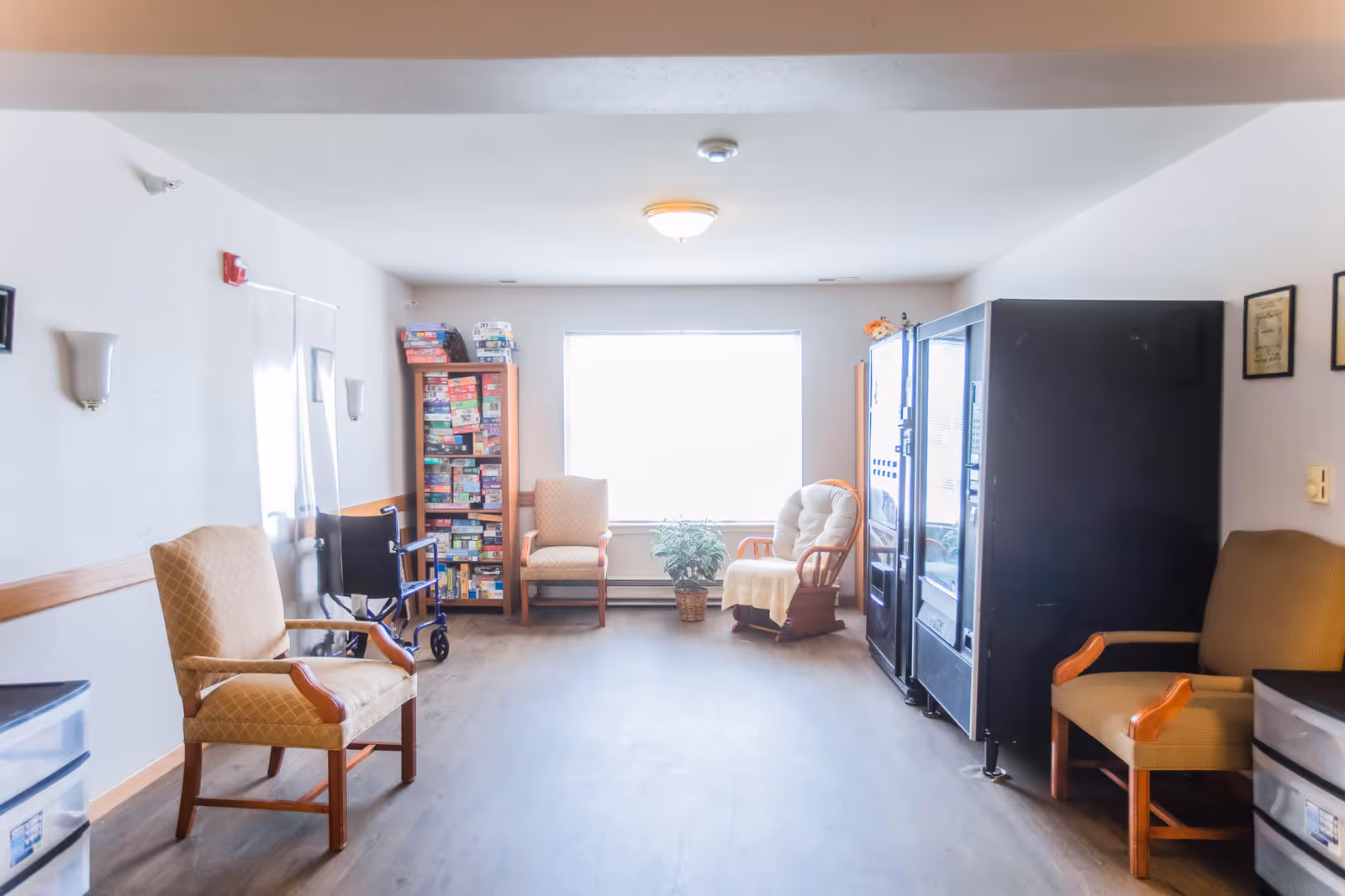 A bright room with several chairs arranged along the walls, a large window letting in natural light, a bookshelf filled with board games, a wheelchair, a potted plant, and two vending machines on the right side.