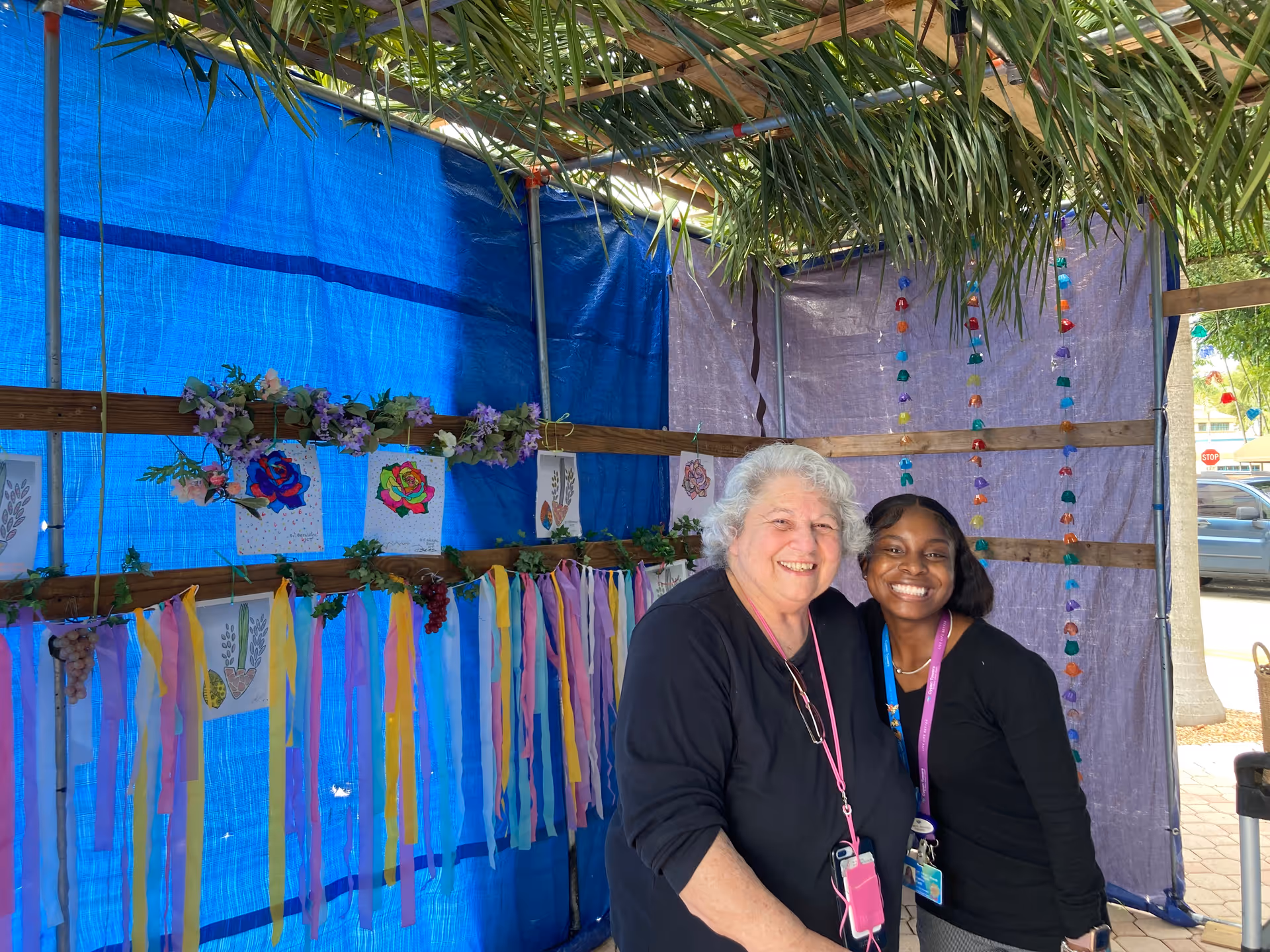 Two women smiling and posing together inside a decorated outdoor structure with blue and purple fabric walls, colorful hanging ribbons, floral decorations, and drawings of flowers and plants. The roof is made of palm leaves, and a car and stop sign are visible outside.