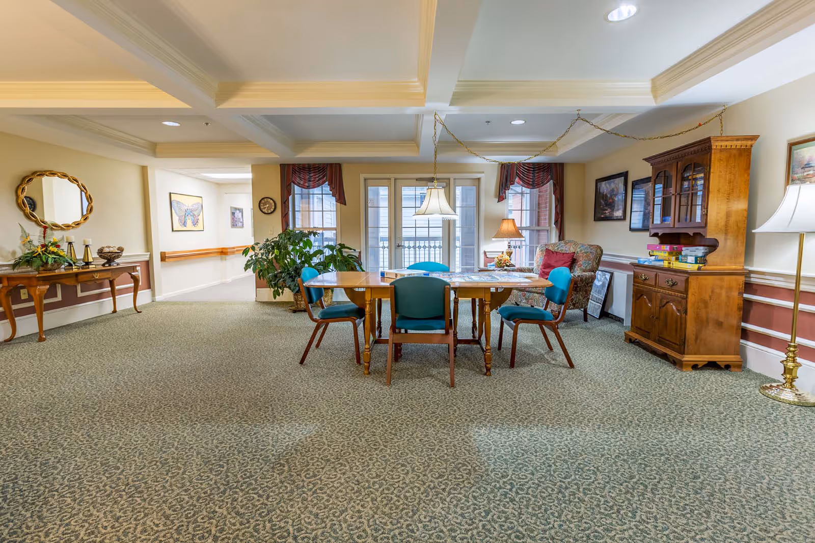 A bright communal dining/activity room with a central wooden table surrounded by teal chairs, cabinets, lamps, and windows.