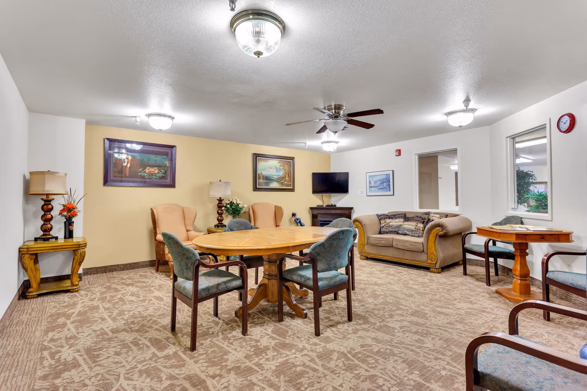 Bright communal sitting room with a round wooden table and chairs, upholstered sofa and armchairs, a TV on a console, and decorative lamps.