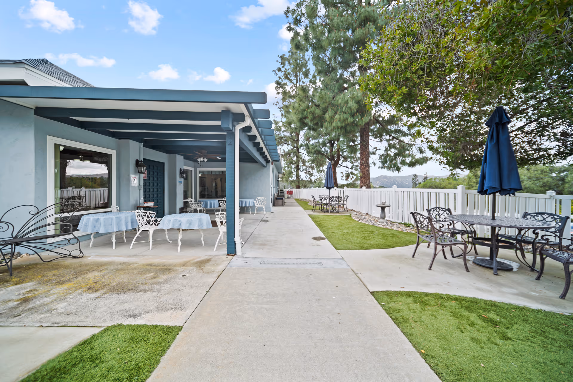 Outdoor patio area at Alpine View Lodge featuring several tables with chairs and umbrellas. The patio is adjacent to a building with a covered seating area that has tables covered with light blue tablecloths. The area is surrounded by a white fence and trees, with a concrete walkway running through the middle.