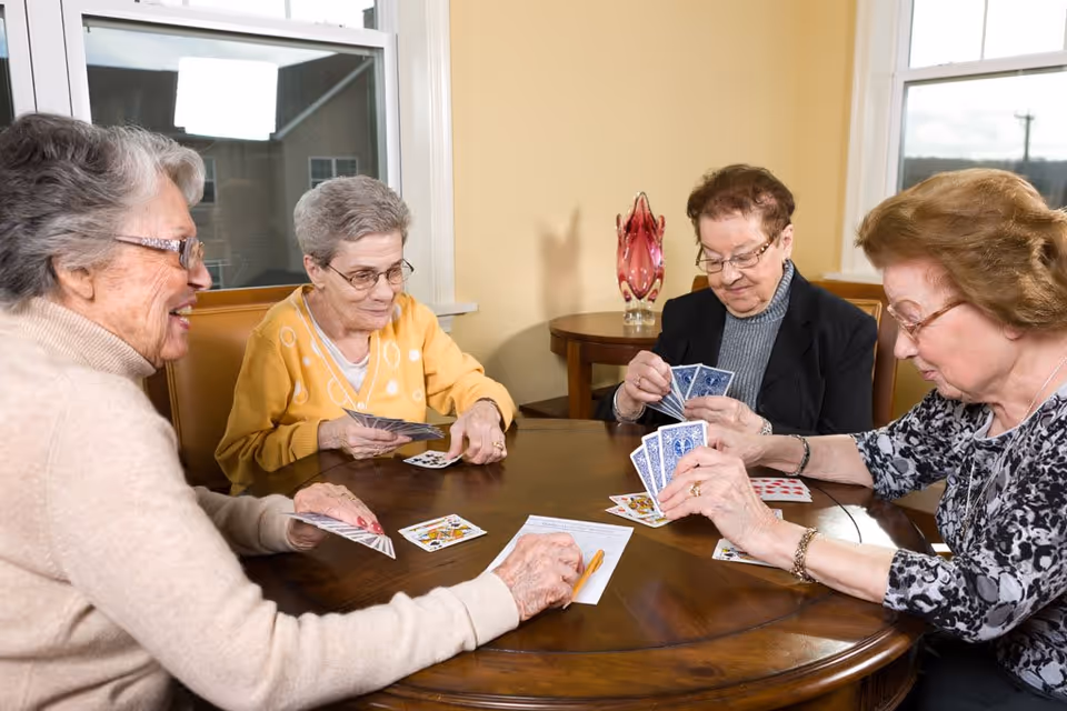 Four elderly women sitting around a wooden table playing cards together in a well-lit room with windows and a decorative glass vase on a side table.