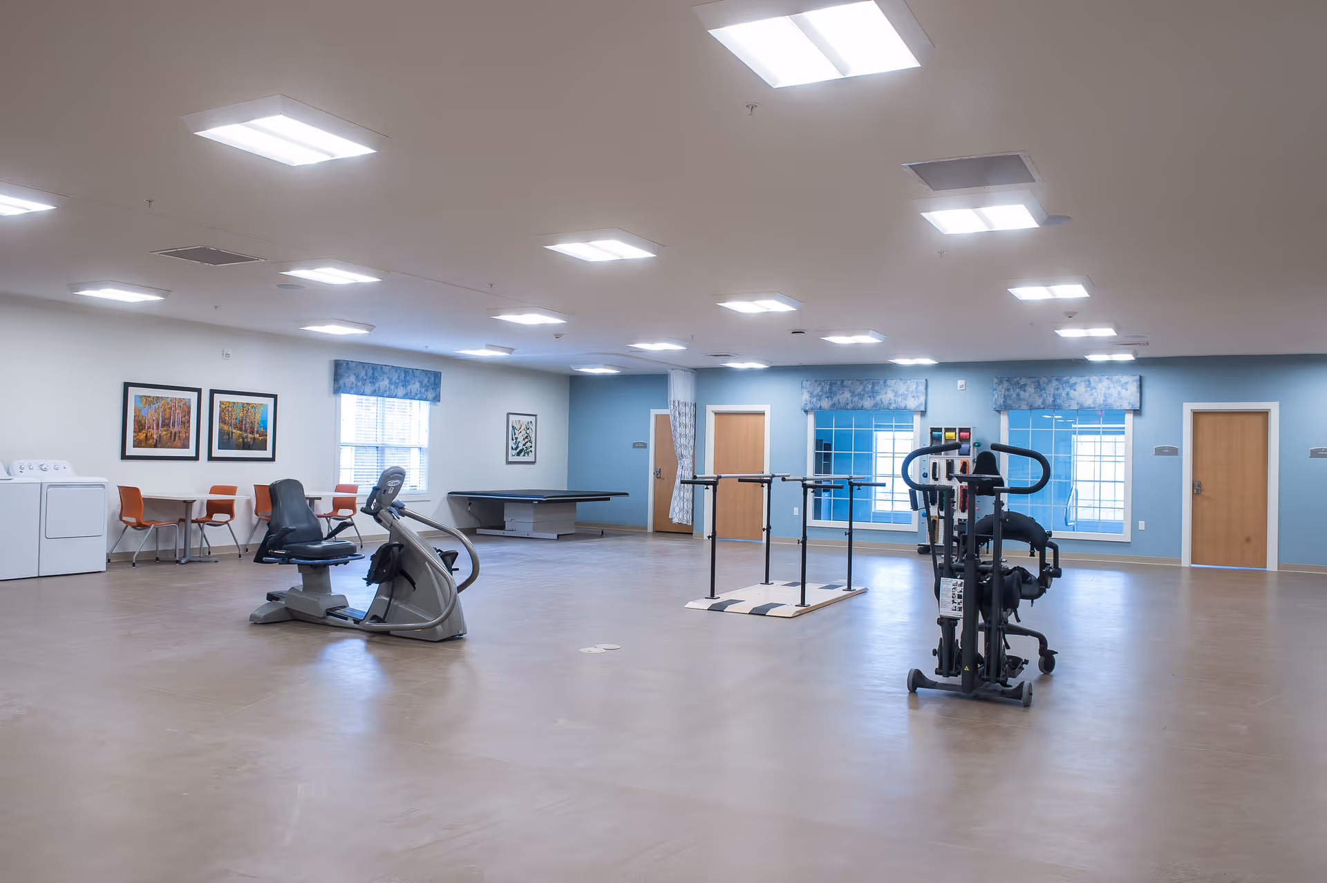 A spacious exercise and activity room in a senior living facility with exercise equipment including a recumbent bike and parallel bars. The room has light blue walls, multiple windows with blue floral valances, framed artwork, and a table with orange chairs near a washer and dryer. The floor is smooth and light brown, and there are several wooden doors along the walls.