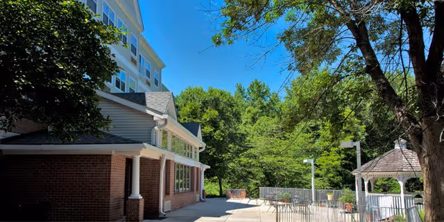 Outdoor patio area of a senior living facility with a brick building on the left, surrounded by green trees under a clear blue sky. The patio includes a white gazebo, railings, and outdoor seating.