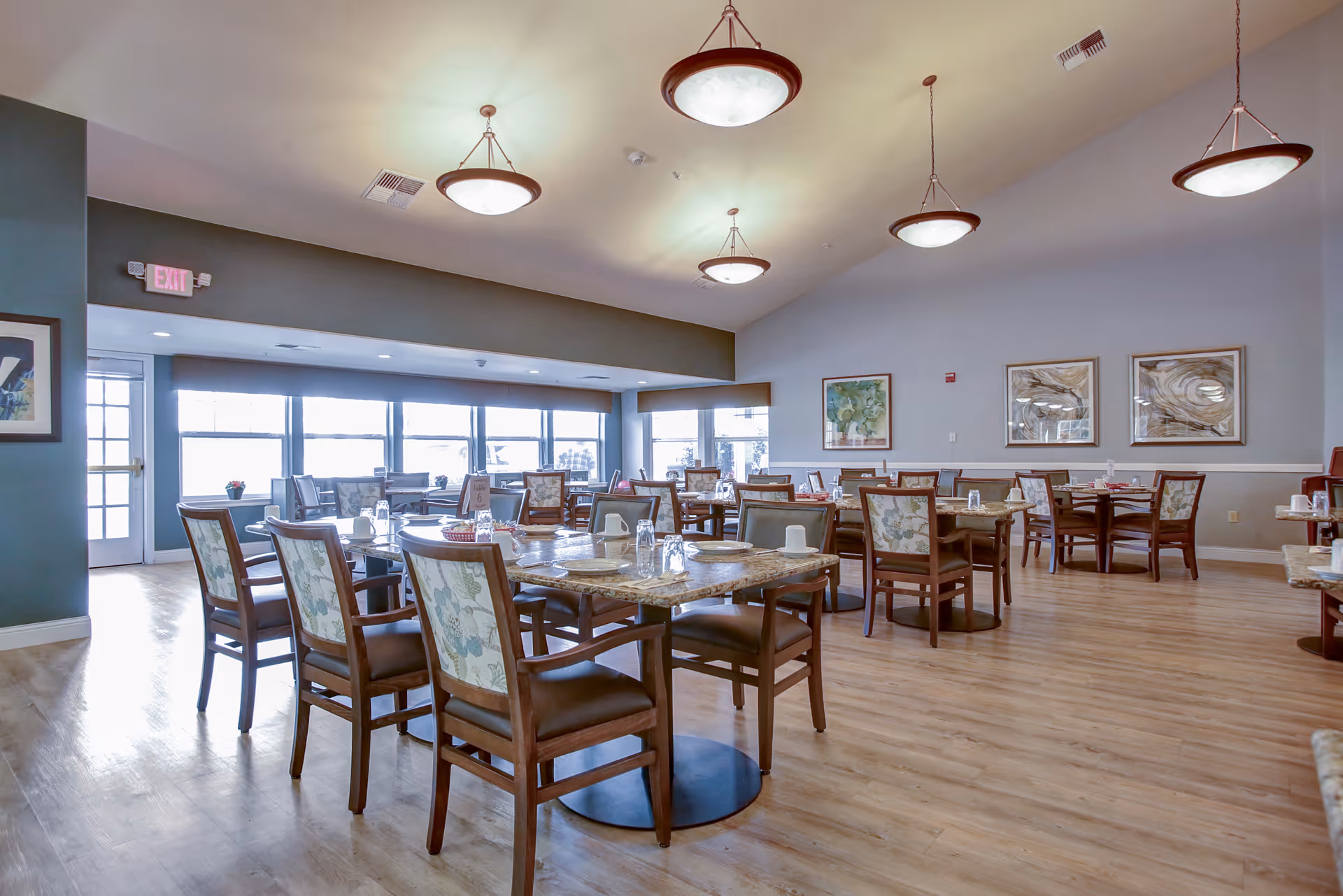 A spacious dining room in a senior living facility with multiple tables and chairs arranged neatly. Each table is set with plates, cups, and glasses. The room has large windows allowing natural light to fill the space, wooden flooring, and several ceiling lights. Artwork is displayed on the walls.