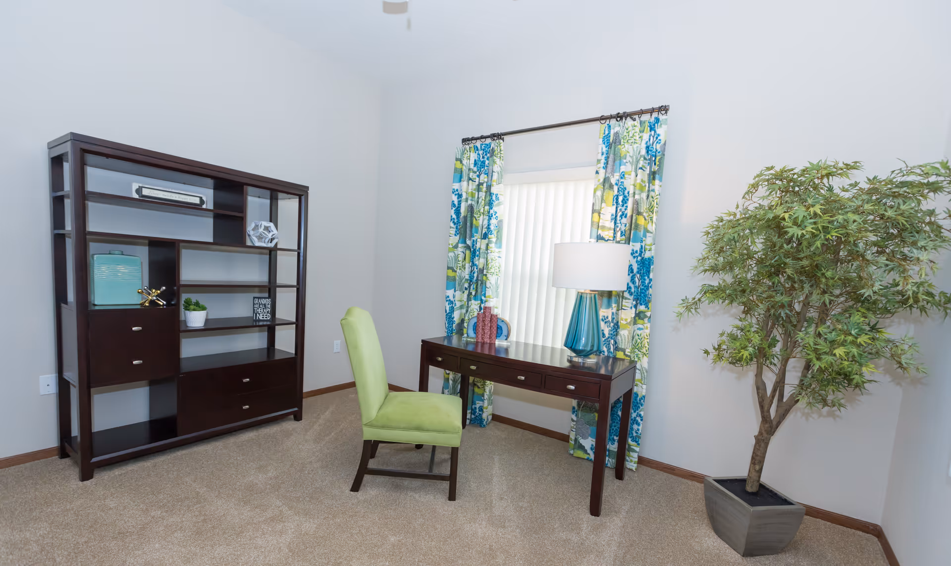 A bright room with beige carpet featuring a dark wooden desk with three drawers, a green upholstered chair, a blue glass table lamp, and decorative items on the desk. Behind the desk is a window with vertical blinds and colorful blue and green patterned curtains. To the left is a dark wooden shelving unit with decorative items and drawers. To the right is a large potted artificial tree with green leaves.
