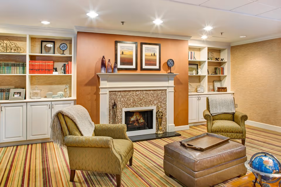 Cozy common room with a fireplace flanked by built-in shelves, two upholstered armchairs, and a leather ottoman.