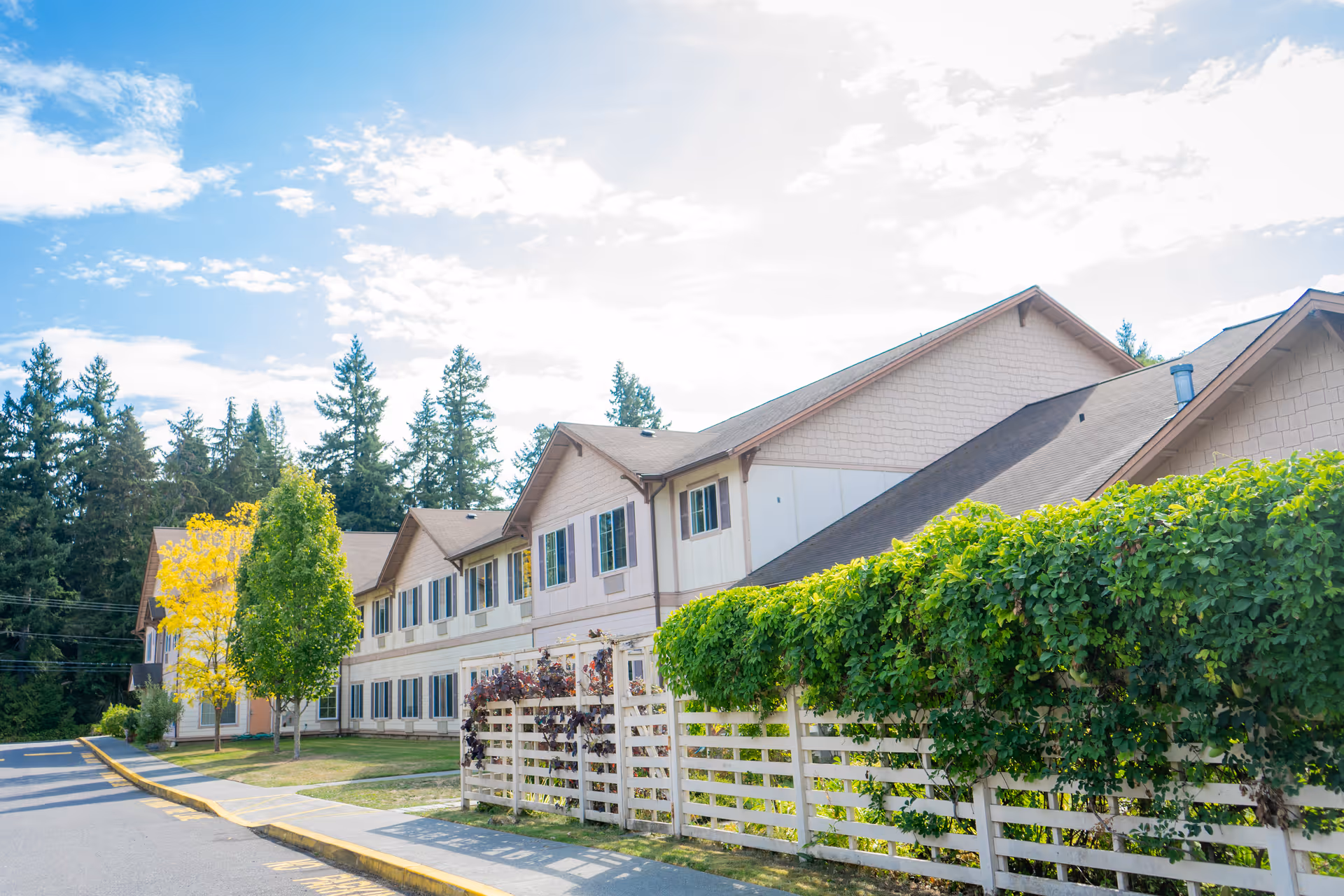 Exterior view of a two-story senior living facility building with beige siding and multiple windows, surrounded by trees and greenery under a partly cloudy sky.