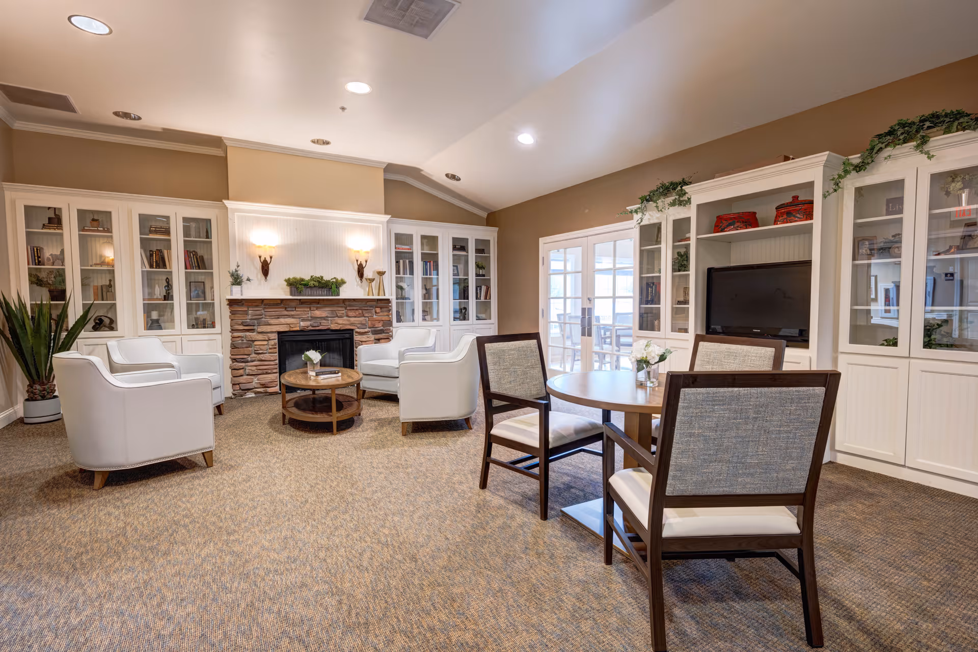 A cozy living room area with beige walls and carpeted floor. The room features a stone fireplace with two wall sconces above it, surrounded by white built-in cabinets filled with books and decorative items. Four white armchairs are arranged around a round wooden coffee table in front of the fireplace. To the right, there is a round wooden table with four chairs, and a flat-screen TV is placed within the built-in cabinetry. The room is well-lit with recessed ceiling lights and has a set of glass double doors leading to another area.