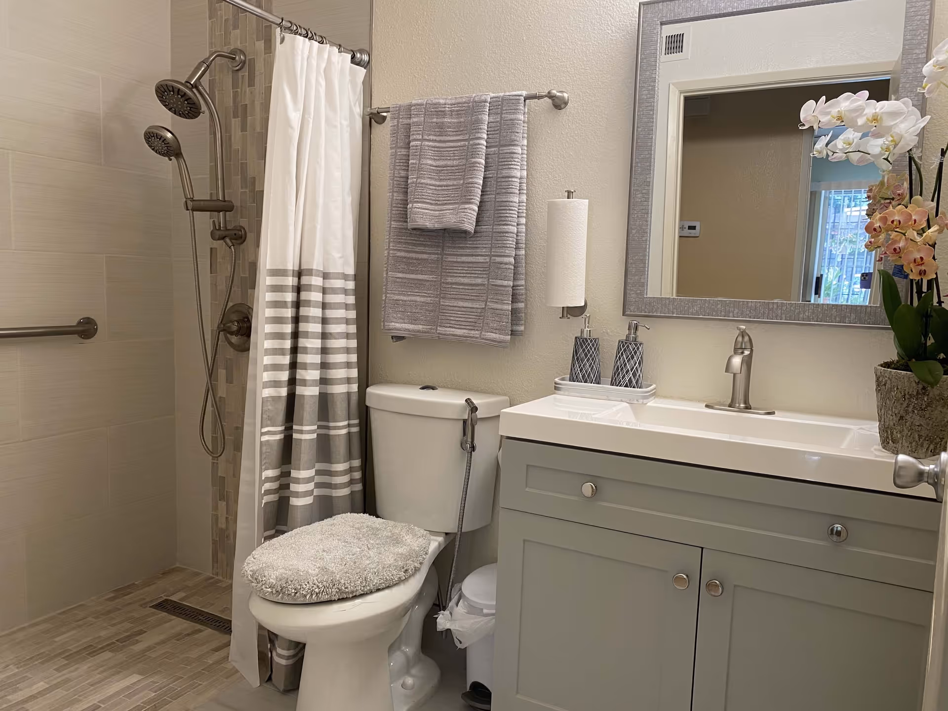 Small modern bathroom with a walk-in shower, toilet, sink vanity with mirror, gray towels, and an orchid on the counter.