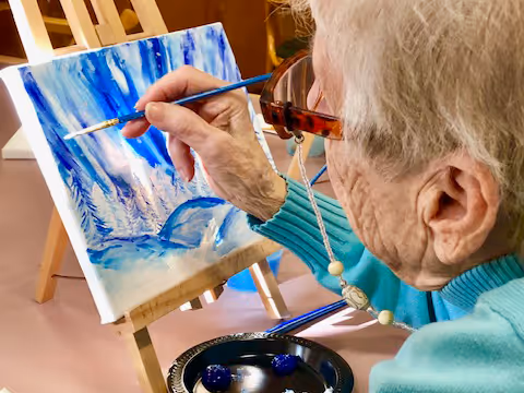 An elderly person wearing glasses and a turquoise sweater is painting a blue and white winter landscape on a small canvas set on an easel. A paint palette with blue paint is visible in front of them.