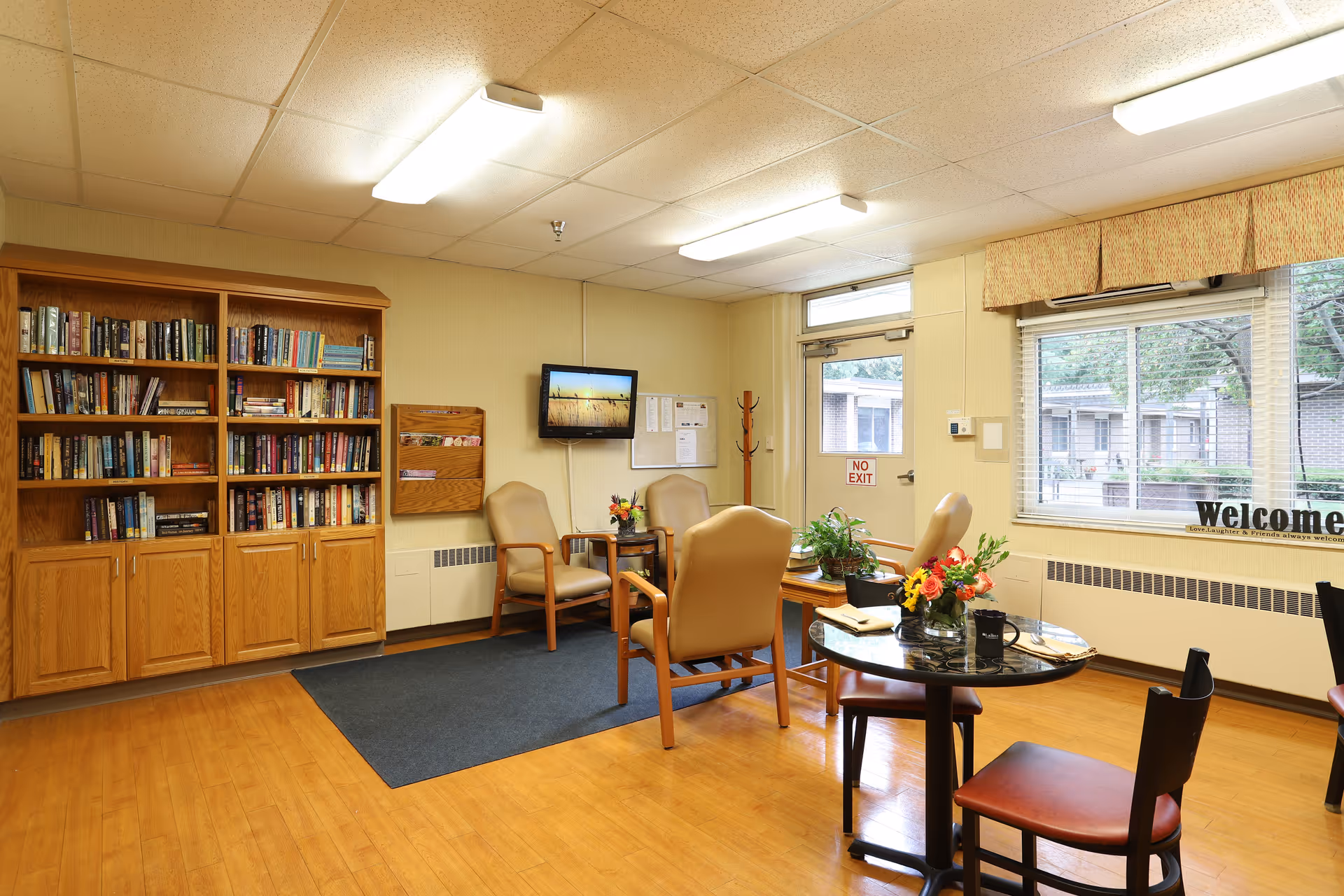 A cozy common area in a senior living facility featuring a wooden bookshelf filled with books, four beige armchairs arranged around a small table with flowers, a round table with chairs and a flower arrangement, a wall-mounted TV, a door with a 'No Exit' sign, and a large window with a 'welcome' sign on the windowsill.