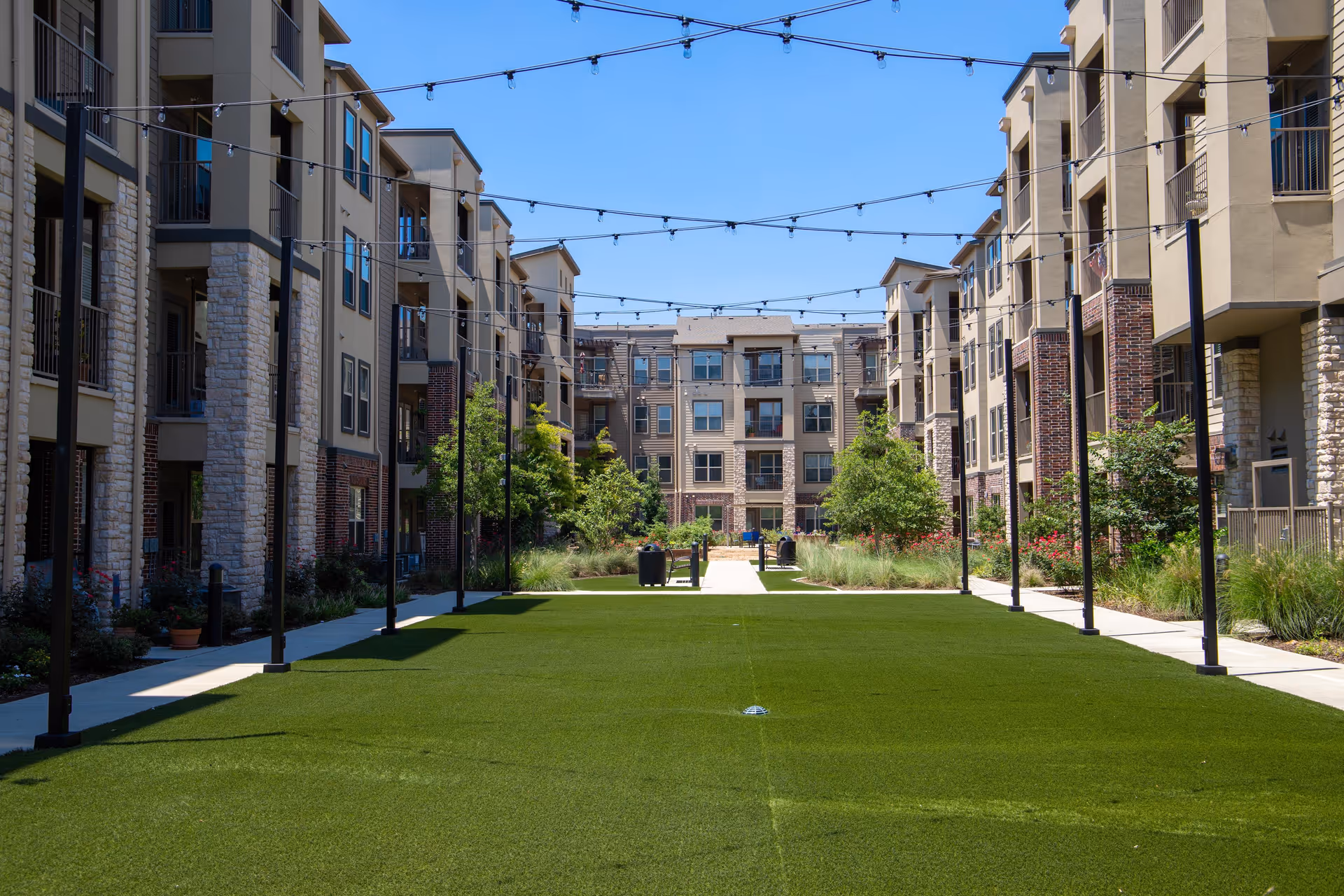 Outdoor courtyard area between multi-story residential buildings with green grass, trees, and string lights overhead under a clear blue sky.