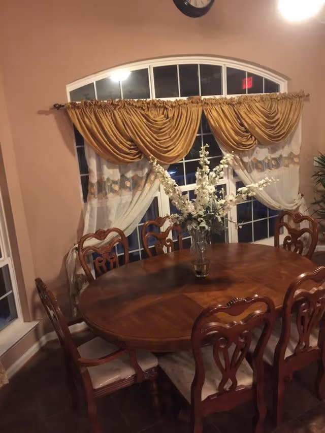 A dining room with a wooden oval table surrounded by six wooden chairs with cushioned seats. A vase with white flowers is placed in the center of the table. Behind the table is a large window with sheer white curtains and gold drapes. The walls are painted a warm beige color, and a clock is visible above the window.