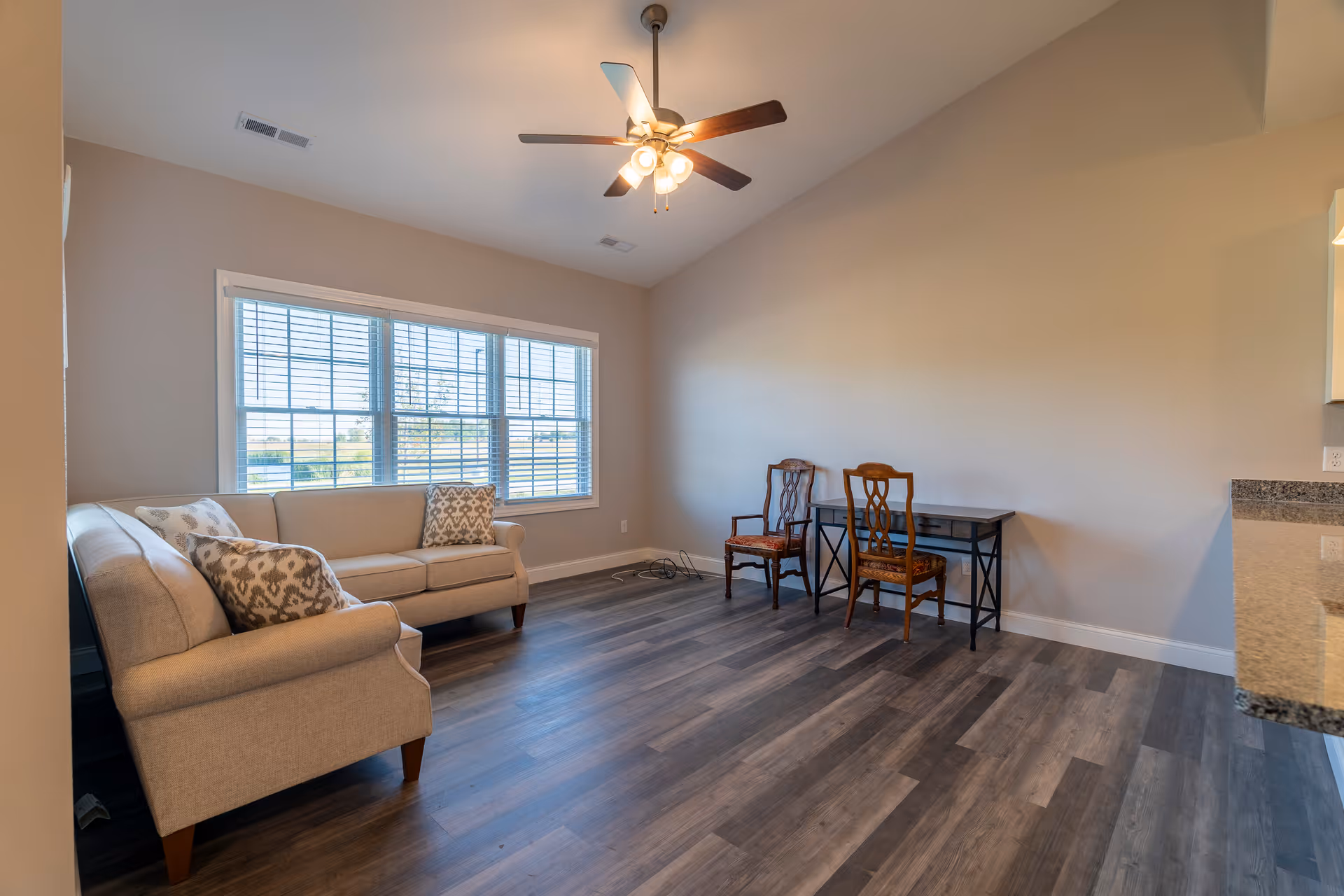 A bright living room with a large window letting in natural light. The room features a beige sofa with patterned cushions on the left side, two wooden chairs with cushions placed in front of a black desk against the right wall, and a ceiling fan with lights overhead. The floor is covered with dark wood laminate, and the walls are painted a light beige color.