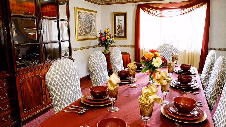 A formal dining room with a long table covered by a red tablecloth, set with brown plates, bowls, and glasses with gold napkins. The chairs have white and gray patterned covers. There is a flower arrangement in the center of the table. The room has a large wooden china cabinet, framed artwork on the walls, and a window with sheer white curtains and red drapes.