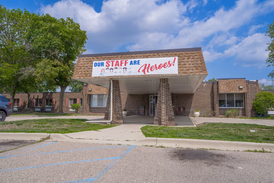 Front exterior view of a single-story brick building with a covered entrance supported by brick pillars. A large banner above the entrance reads 'OUR STAFF ARE Heroes!' with illustrations of people wearing masks. There are trees and parked cars visible around the building under a partly cloudy sky.