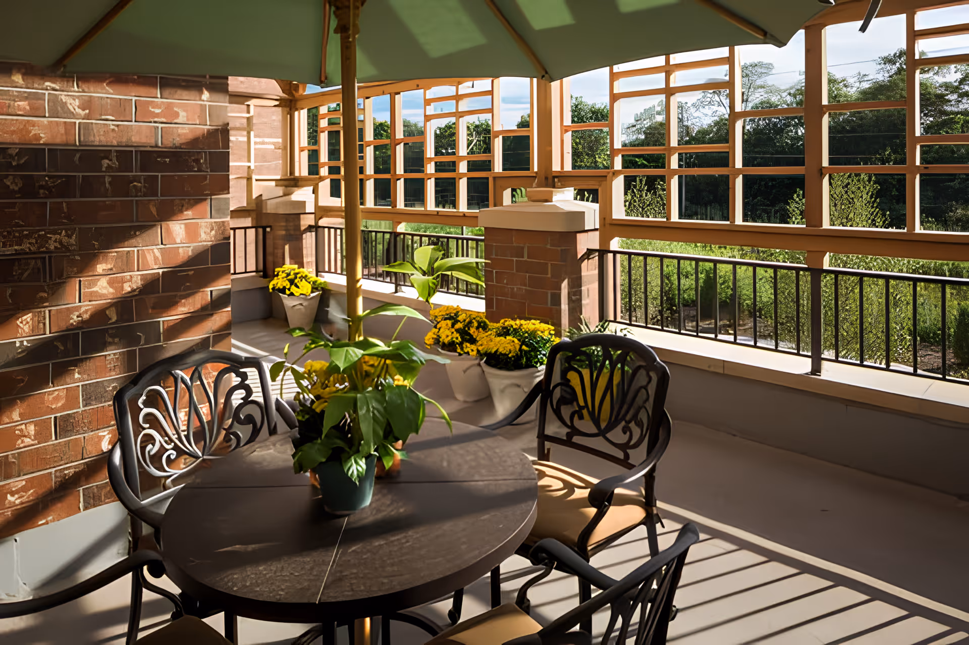A shaded outdoor patio area with a round table and four decorative metal chairs. The table has a potted plant with green leaves. The patio is enclosed with brick pillars and wooden lattice panels, with yellow flowers in pots placed along the edges. Greenery and trees are visible outside the patio.
