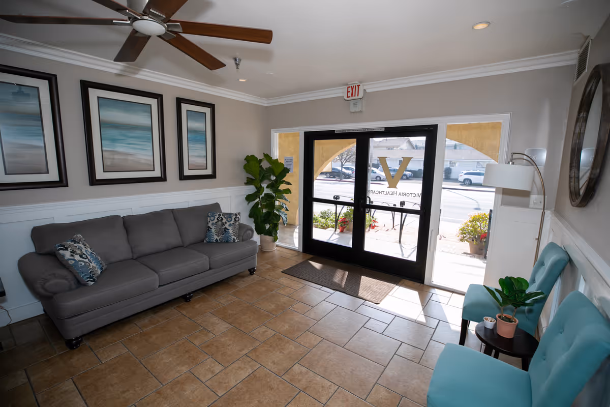A bright and welcoming lobby area with a gray couch adorned with patterned pillows on the left, two turquoise chairs with a small round table holding potted plants on the right, a ceiling fan, three framed ocean-themed paintings on the wall, and glass double doors with the Victoria Healthcare logo visible through them.