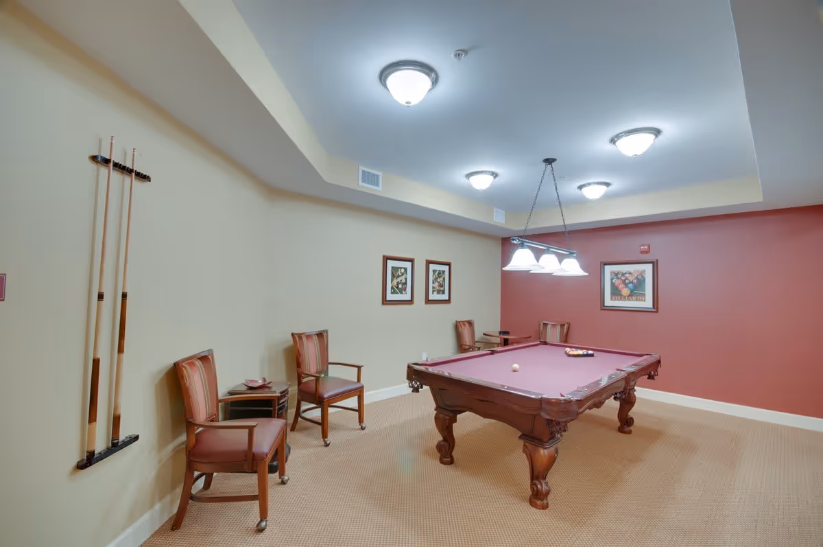 A recreational room with a pool table in the center, surrounded by four wooden chairs with cushions. The walls are painted beige and red, with framed pictures hanging on them. Pool cues are mounted on the beige wall, and ceiling lights illuminate the room.