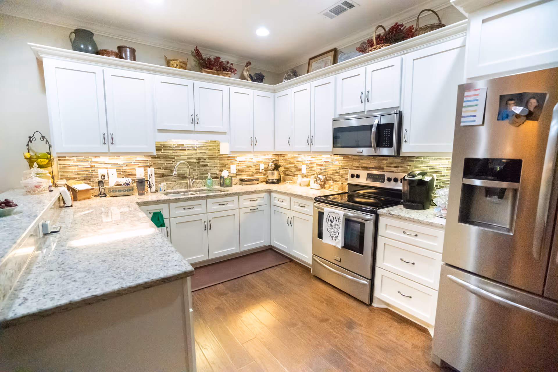A modern kitchen with white cabinets, granite countertops, and a tiled backsplash. The kitchen features a stainless steel refrigerator, oven, and microwave. There are various kitchen items on the counters, including a coffee maker, a fruit basket, and decorative items on top of the cabinets. The floor is wooden, and the kitchen is well-lit with under-cabinet lighting.