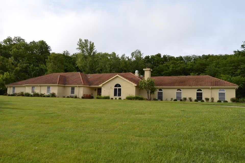 Single-story beige building with a red-tiled roof surrounded by a large green lawn and trees in the background under a cloudy sky.