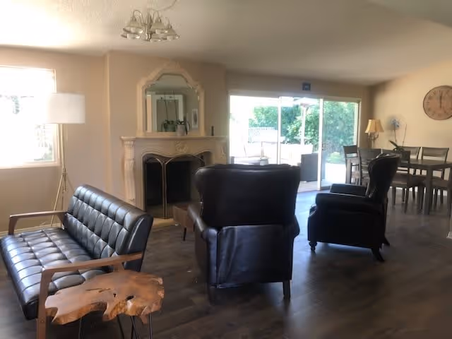 A cozy living room area with a black leather sofa and two black leather armchairs arranged around a fireplace with a decorative mirror above it. There is a wooden side table with a natural edge next to the sofa, a floor lamp near a window, and a dining table with chairs in the background near sliding glass doors that open to an outdoor patio.