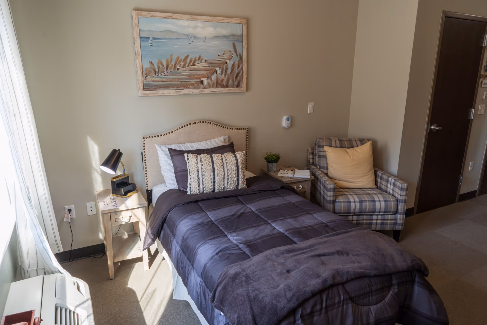 A cozy bedroom with a single bed covered in a dark comforter and multiple pillows. Next to the bed is a small wooden nightstand with a black lamp and some items on it. A plaid armchair with a yellow cushion is placed beside the nightstand. A framed painting of a pier and water hangs on the wall above the bed. Light filters in through a window with sheer curtains on the left side of the room.