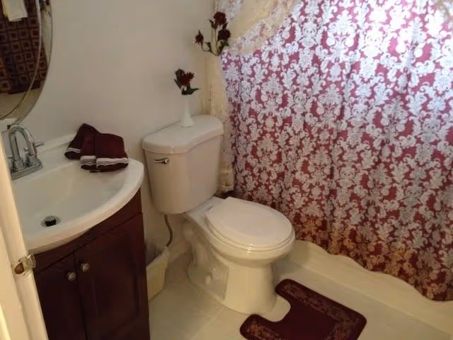 A small bathroom featuring a white toilet with a white cushioned seat, a dark wood vanity with a white sink and silver faucet, a round mirror above the sink, and a shower with a red and white patterned curtain. There are burgundy towels on the sink and a matching burgundy bath mat on the floor. A small vase with dark red flowers is placed on top of the toilet tank.