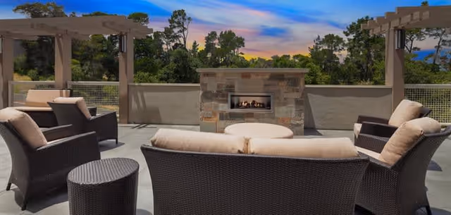 Outdoor patio area with cushioned wicker chairs and sofas arranged around a stone fireplace, with pergolas on either side and trees in the background under a colorful evening sky.
