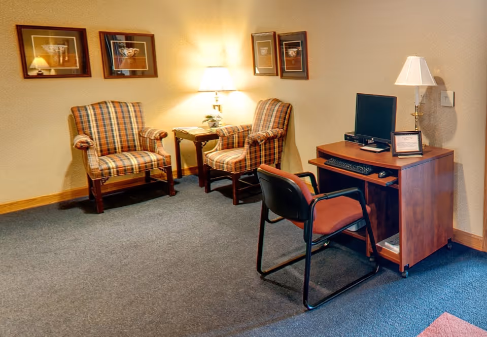 A cozy interior room with two plaid upholstered armchairs and a small wooden side table with a lamp and a plant. On the right side, there is a wooden desk with a computer monitor, keyboard, mouse, and a framed certificate, accompanied by a red cushioned chair. The walls are decorated with framed pictures and the floor is carpeted.