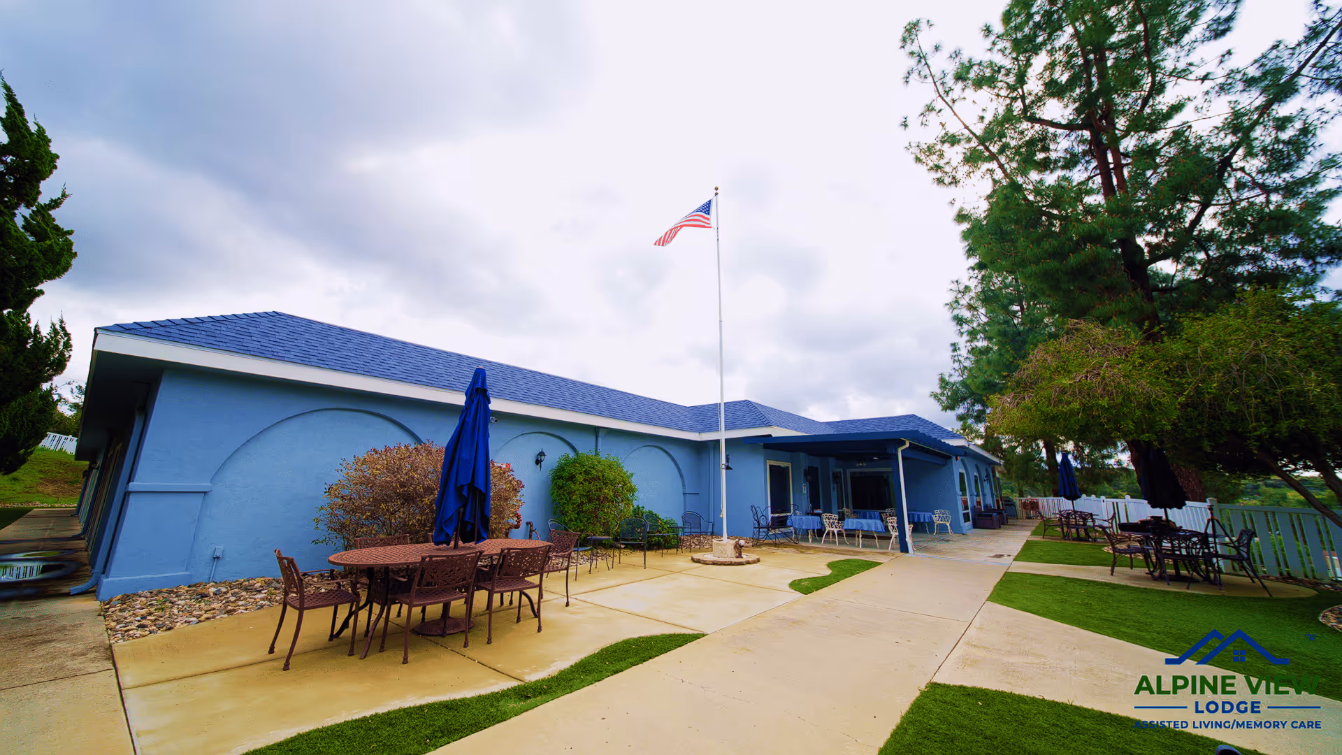 Outdoor patio area at Alpine View Lodge featuring multiple metal tables and chairs with blue umbrellas, a concrete walkway, green artificial grass patches, and an American flag on a flagpole in front of a blue building under a cloudy sky.